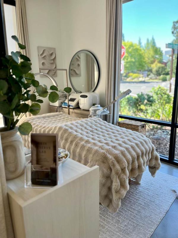 Treatment bed beside a floor-to-ceiling window, with a round mirror and skincare devices behind it.